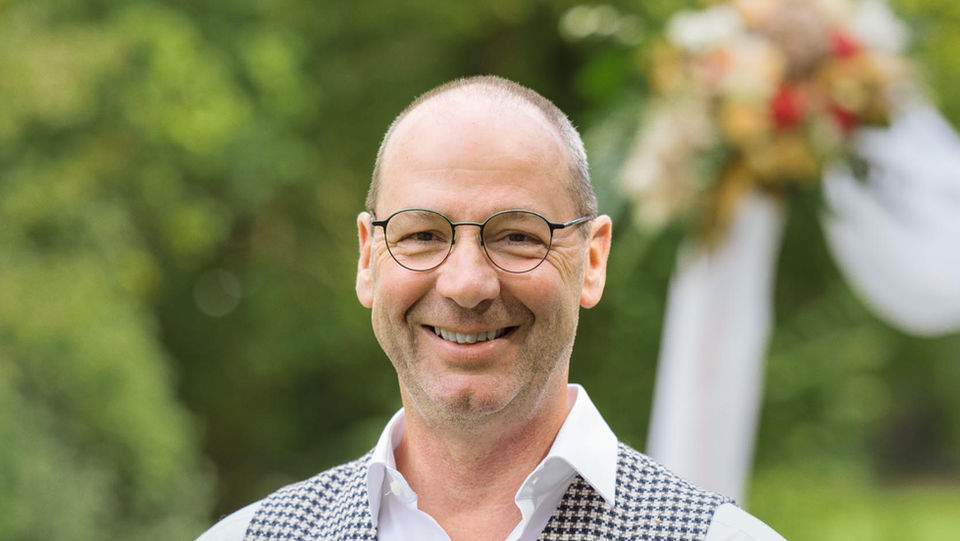 Trauredner Jochen Andreae hält ein Buch, lächelt in die Kamera, Hintergrund im Freien.