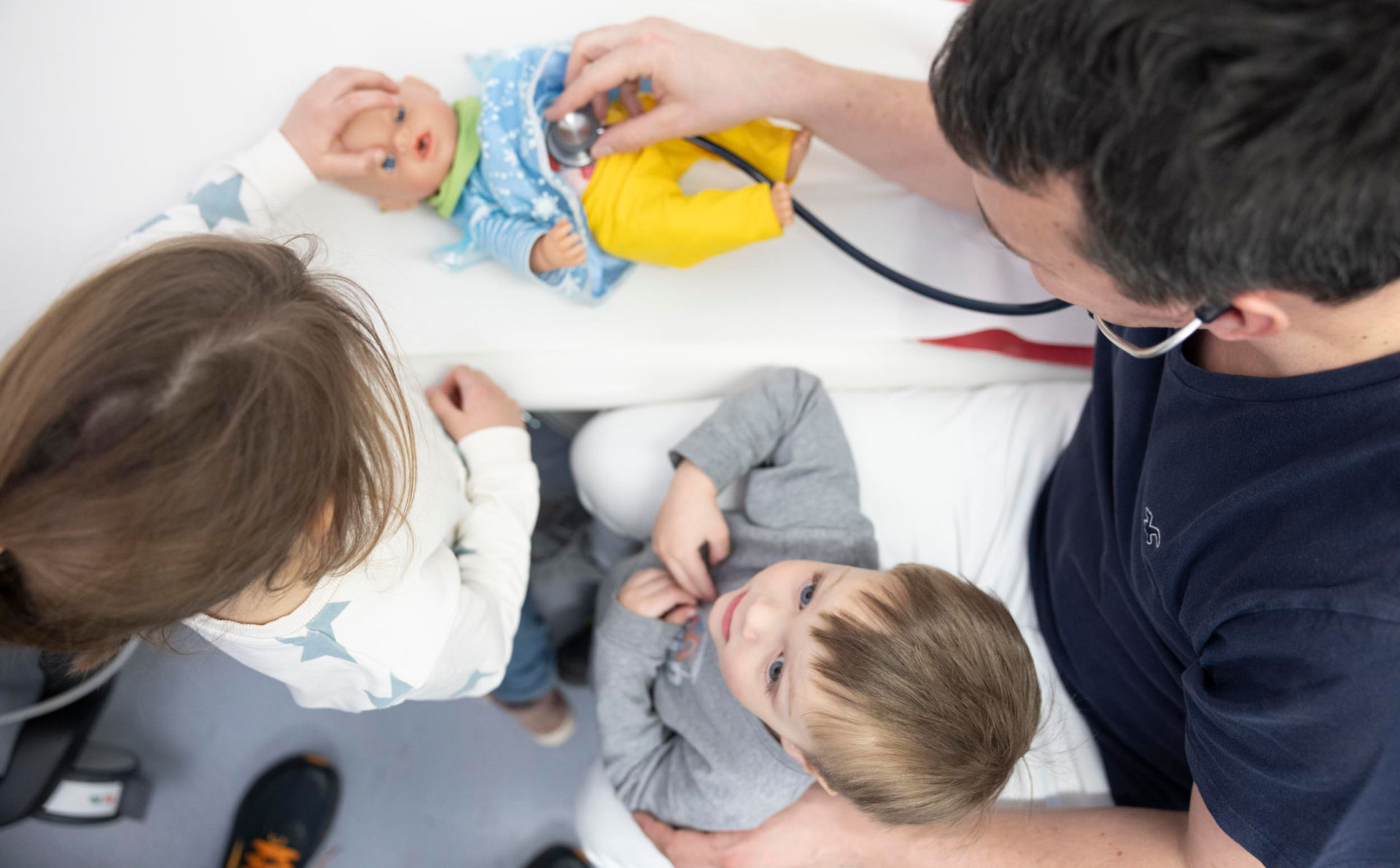 Dr. Med. Linsenmeyer zeigt einem kleinen Kind an dessen Puppe, wie das Abhören mit dem Stethoskop funktioniert.