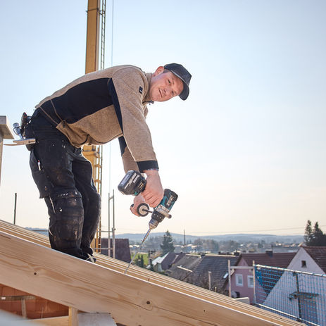 Fröhlicher Bauarbeiter setzt einen Nagel mithilfe eines Bohrers in einen Holzbalken von dem Dachstuhl