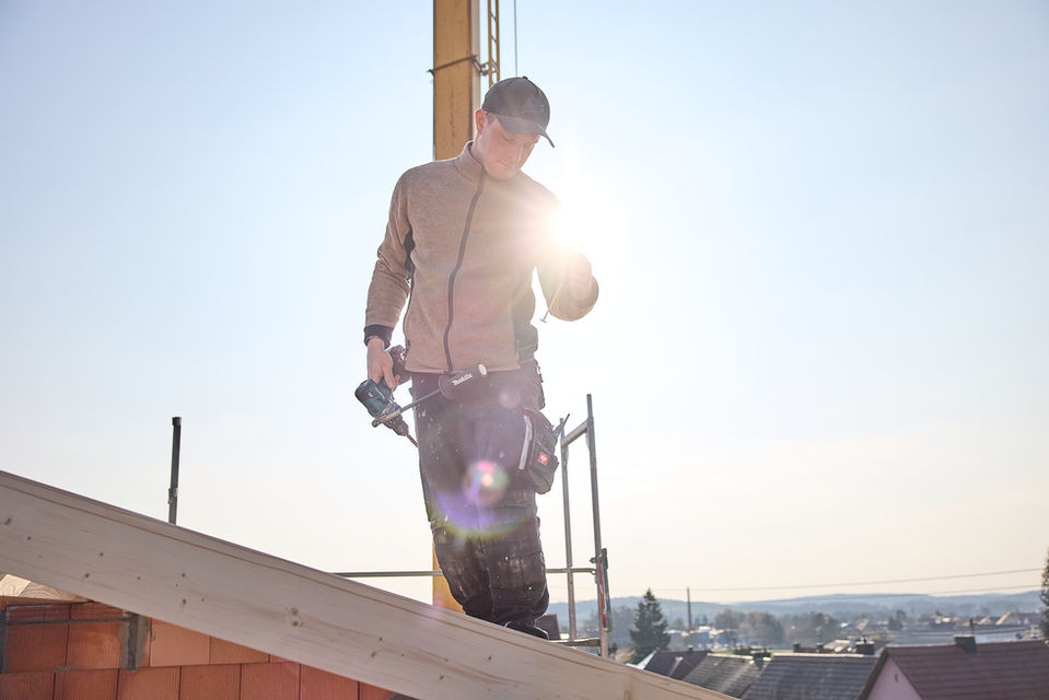Ein Bauarbeiter steht im Dachstuhl. Hinter ihm scheint die Sonne und man sieht einen Kran.