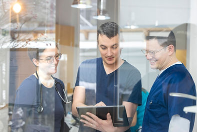 Die drei Ärzte der Gemeinschaftspraxis stehen hinter einer Glasscheibe und besprechen Akten auf einem Tablet.