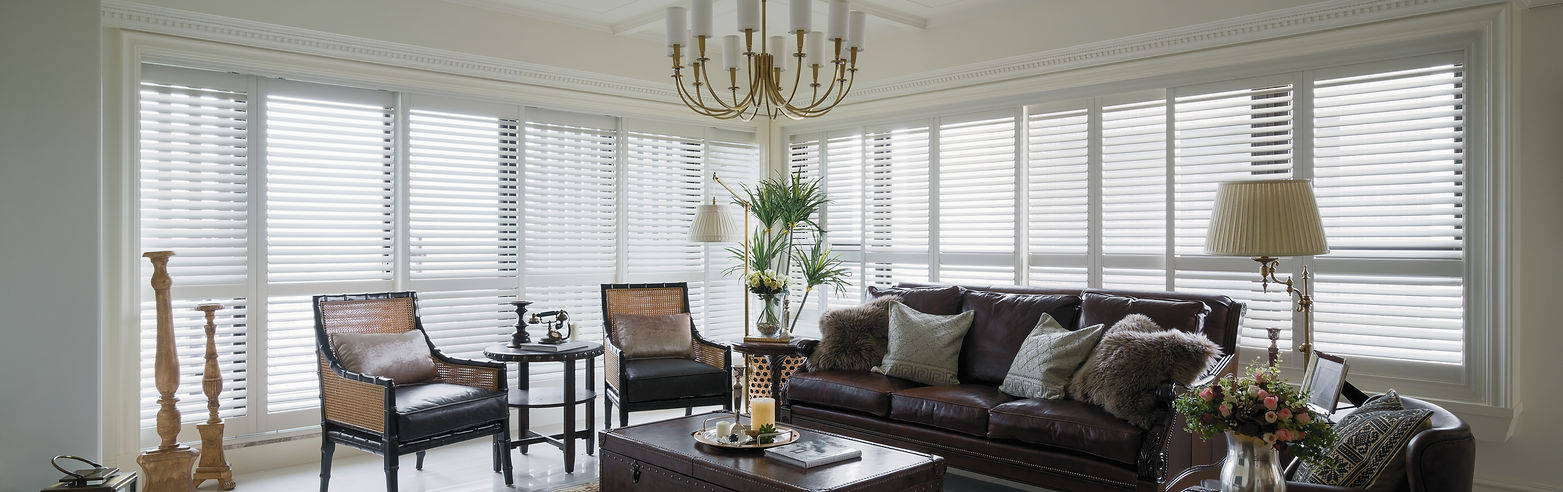 White Norman Brightwood plantation shutters wrap two walls of a sunroom with a dark leather sofa, cane-back armchairs and a brass chandelier.