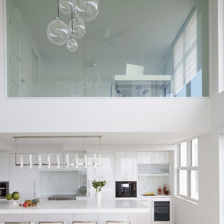 Two-story white kitchen with glass-railed loft, globe chandelier, and glossy modern cabinetry.