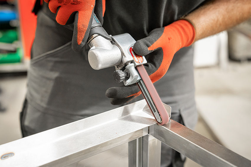 Person wearing orange gloves uses a power tool on a metal frame. The setting is an industrial workshop. The tool features a visible red label.