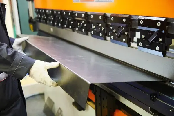 Person in gloves operates an orange metal press brake, bending a steel sheet. Industrial setting with focus on precision machinery.