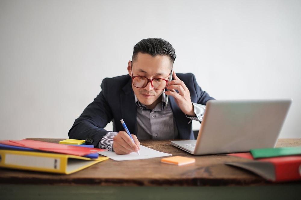 photo of man in blue suit jacket striped shirt and eyeglasses talking on the phone while sitting at a table with his laptop