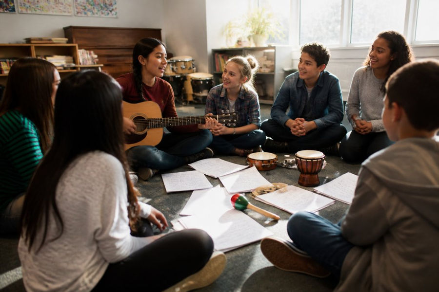 A diverse group of students sitting in a circle in a bright classroom, one student holding a guitar and leading a song while others listen and smile, musical instruments and handwritten lyrics scattered on the floor