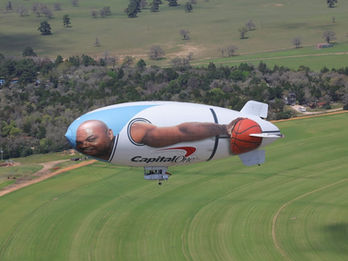 Magic Johnson Capital One promotional blimp and commercial airship flying low over green fields for targeted outdoor event aerial advertising.