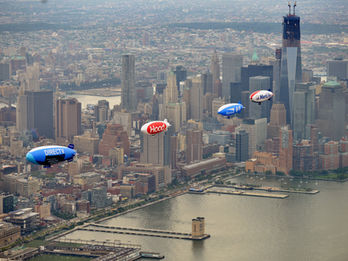 Fleet of commercial advertising blimps and promotional airships flying over a major city skyline and river for metropolitan marketing.