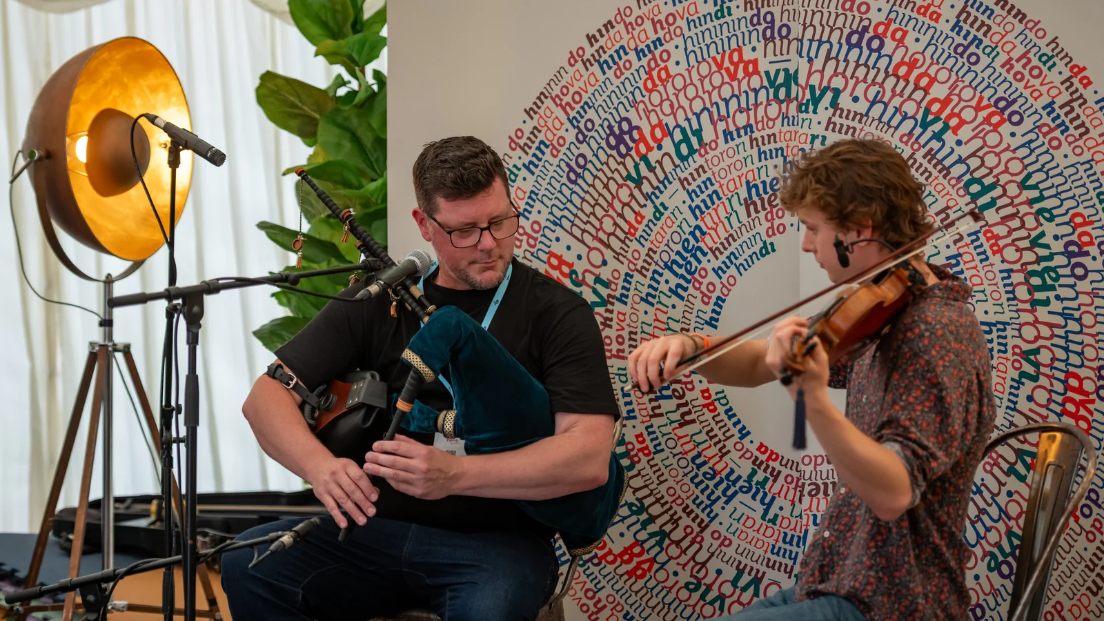 Donald playing smallpipes and Roo playing fiddle, beside a golden stage lamp at Piping Live.