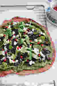 A closeup of the toppings on the beetroot crust pizza, arugula pesto, fresh arugula, ricotta cheese and pomegranate seeds.