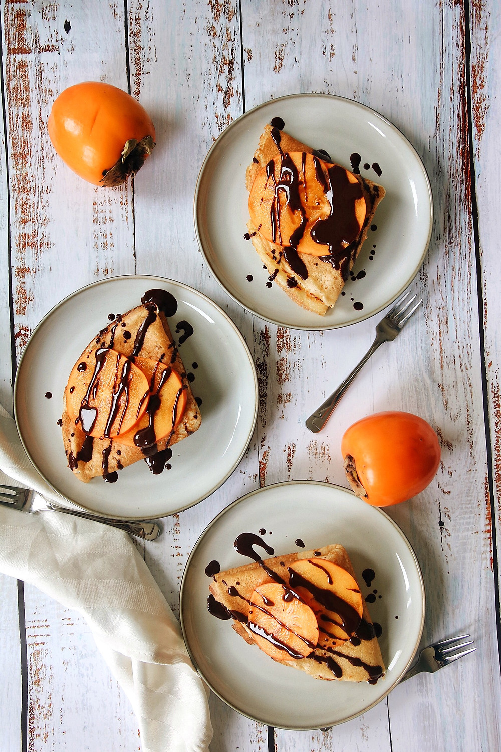 Three plates of gluten free crepes filled with a walnut and rum filling, topped with fresh persimmons and dark chocolate.