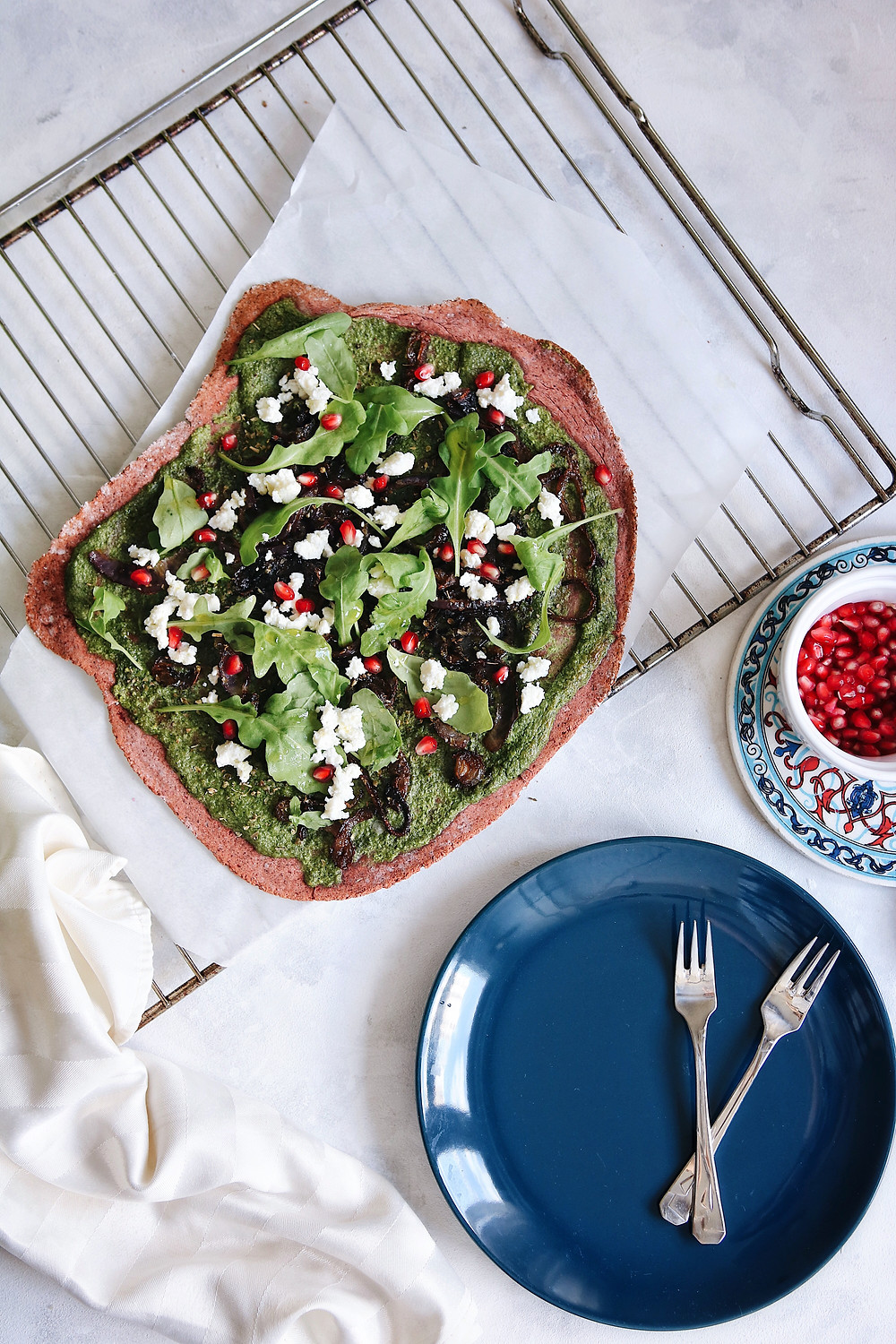 A beetroot crust pizza on a white backround, a cooling rack, blue plate and pomegranate seeds on the side.