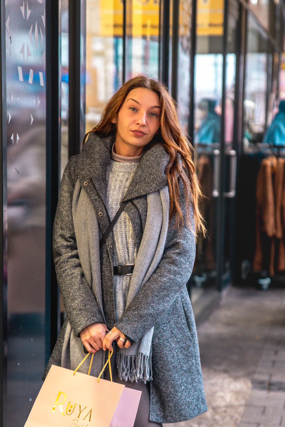 Person in grauem Mantel, Schal und Strickpullover steht vor einer gläsernen Storefront und hält eine Einkaufstasche; im Hintergrund spiegeln sich Schaufensterpuppen und Lichter.