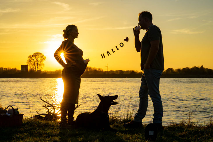 Schwangere Person und Partner stehen bei Sonnenuntergang am Fluss, verbunden durch Schnurtelefon mit „HALLO“-Botschaft.