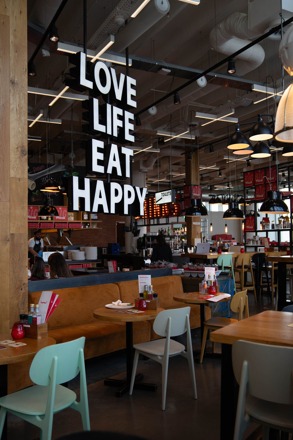 Das Innere eines modernen Restaurants mit bunten Sitzgelegenheiten, warmem Licht und lebendiger Atmosphäre. Über dem Raum hängt in großen Leuchtbuchstaben der Schriftzug „LOVE LIFE EAT HAPPY“. Im Hintergrund befindet sich eine Bar mit Flaschen und Gläsern, sowie ein Schild mit der Aufschrift „TOILETS“. Gäste und Personal beleben die Szene.