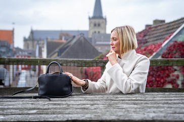 Frau mit hellem Mantel sitzt an Holztisch mit Handtasche vor Altstadt-Kulisse und herbstlichem Laub.