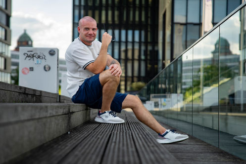 Mann mit weißem Shirt und Shorts sitzt auf Holzstufen vor moderner Stadtkulisse mit Glasfassaden.