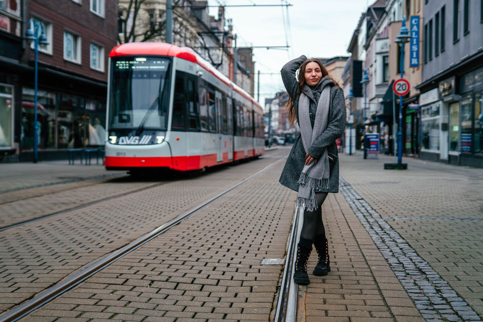 Person in grauem Mantel und Schal steht auf Straßenbahngleisen und posiert, während eine Tram im Hintergrund heranfährt; umgeben von städtischen Gebäuden, Geschäften und Verkehrselementen.
