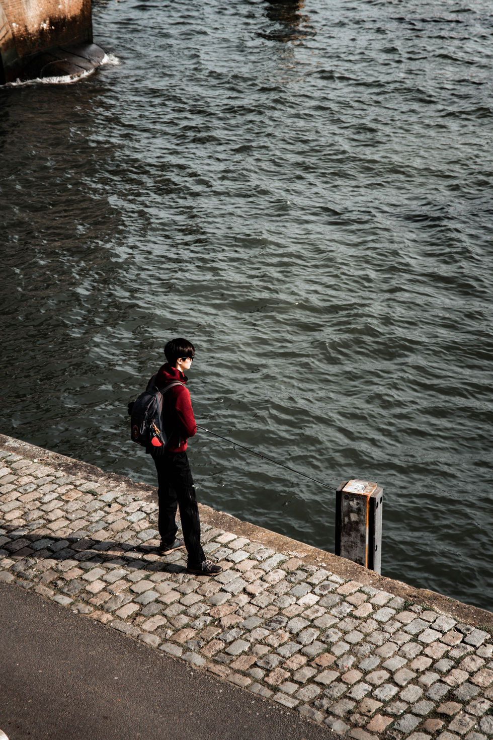 Eine Person in roter Jacke und mit Rucksack steht auf einem Kopfsteinpflasterweg am Wasser und angelt. Das leicht bewegte Wasser trifft auf eine Beton- und Metallstruktur am Ufer. Die Szene vermittelt Ruhe und Konzentration inmitten urbaner Natur.