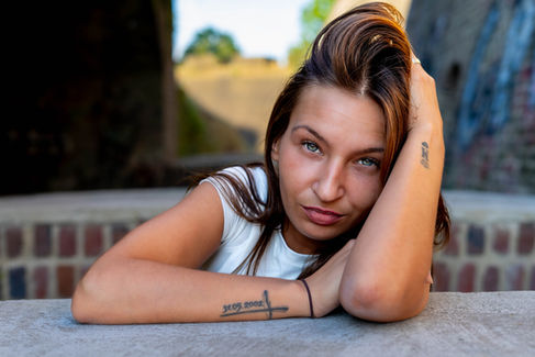 Person mit Tattoos lehnt sich nachdenklich auf Betonfläche vor Backsteinwand und Bäumen.