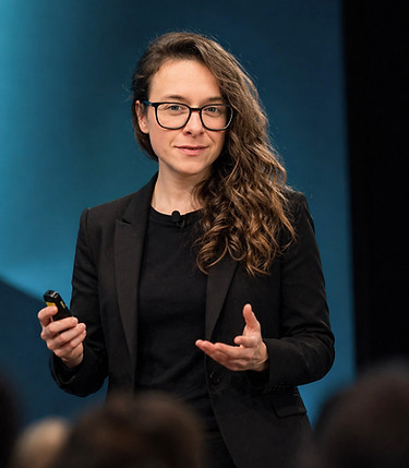 Laurence Paquette, a woman with long wavy brown hair and black-rimmed glasses stands on a stage, speaking and gesturing with one hand open while holding a presentation clicker in the other. She is wearing an all-black outfit against a softly lit teal blue background, with the blurred heads of an audience visible in the foreground.