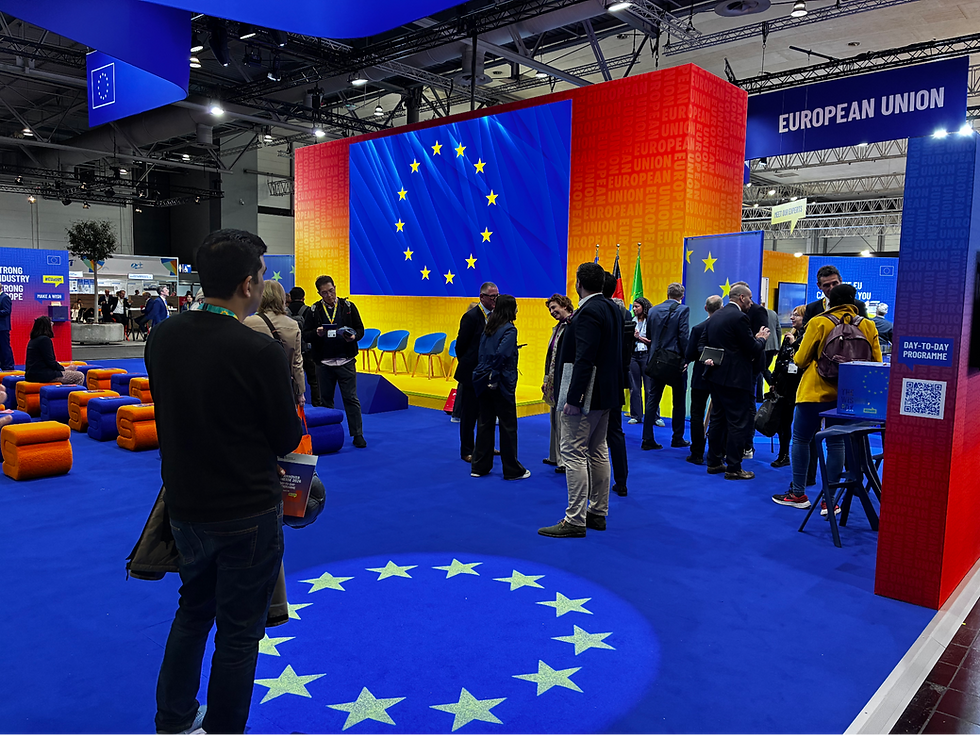 People gather in a vibrant EU-themed booth with a blue and yellow color scheme, featuring EU flags and signage. The mood is busy and engaged.