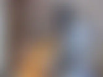 A young girl in a yellow sweater interacts with a silver robot in a cozy kitchen. She touches its chest screen, with shelves in the background.