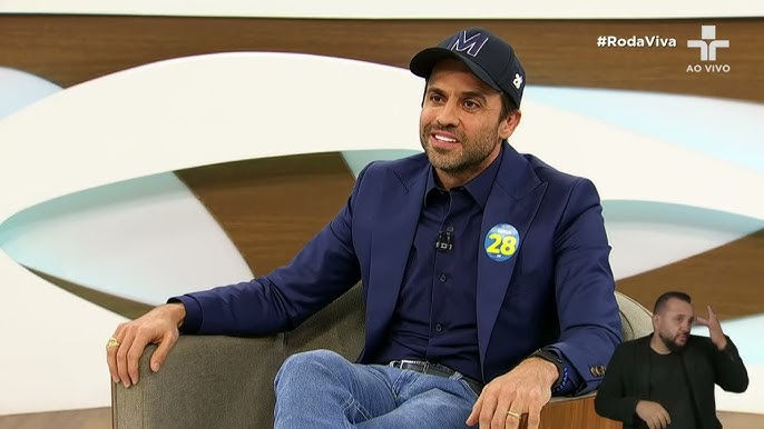 Pablo Marçal em uma entrevista no programa Roda Viva, vestindo um boné com a letra "M" e um adesivo de campanha, aplicando técnicas de marketing digital baseadas em vídeos curtos e polêmicas calculadas.