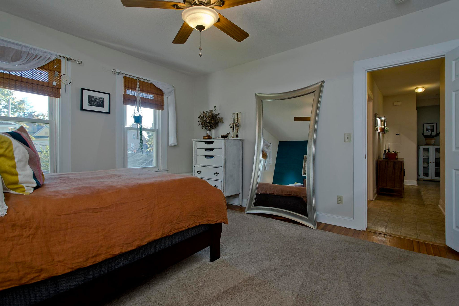 This is a good sized bedroom, with a door leading to the hallway, a large mirror against the wall, a dresser in the corner. The bed is next to the wall with two windows. The walls are light tan, and trim is white. 