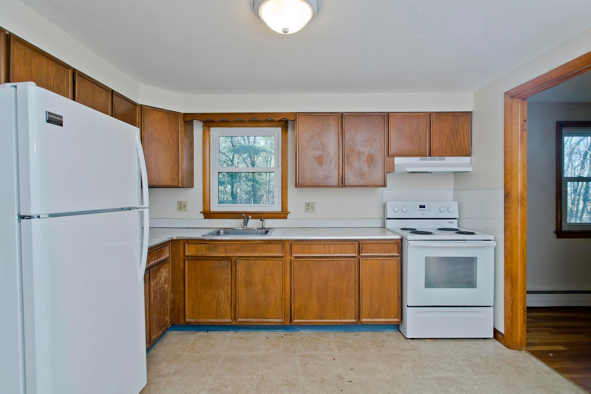 There is a slightly dated kitchen with wooden cabinets, white countertops and white appliances. There is a small window above the sink, and to the right a doorway to another room.