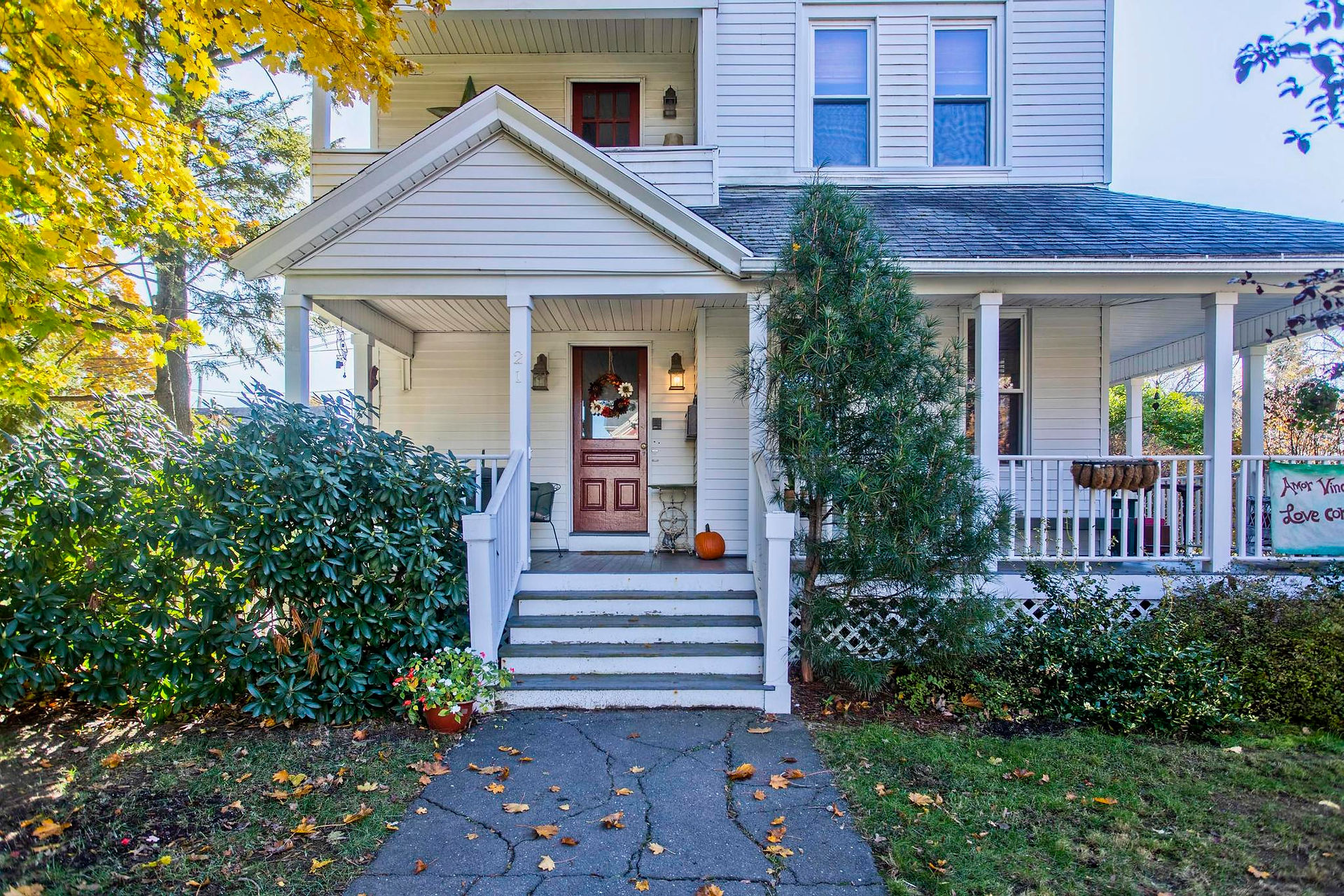 There is a white 3 story house, with a small tree in the front right yard, and a larger tree to the left. A sidewalk leads to the front door.