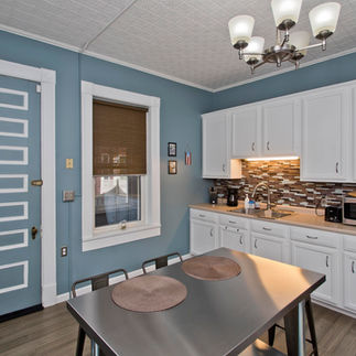 The ceilings in this kitchen are tall, and white. The walls are a medium blue color and cabinets are white, with stone looking backsplash. The floor is grey.