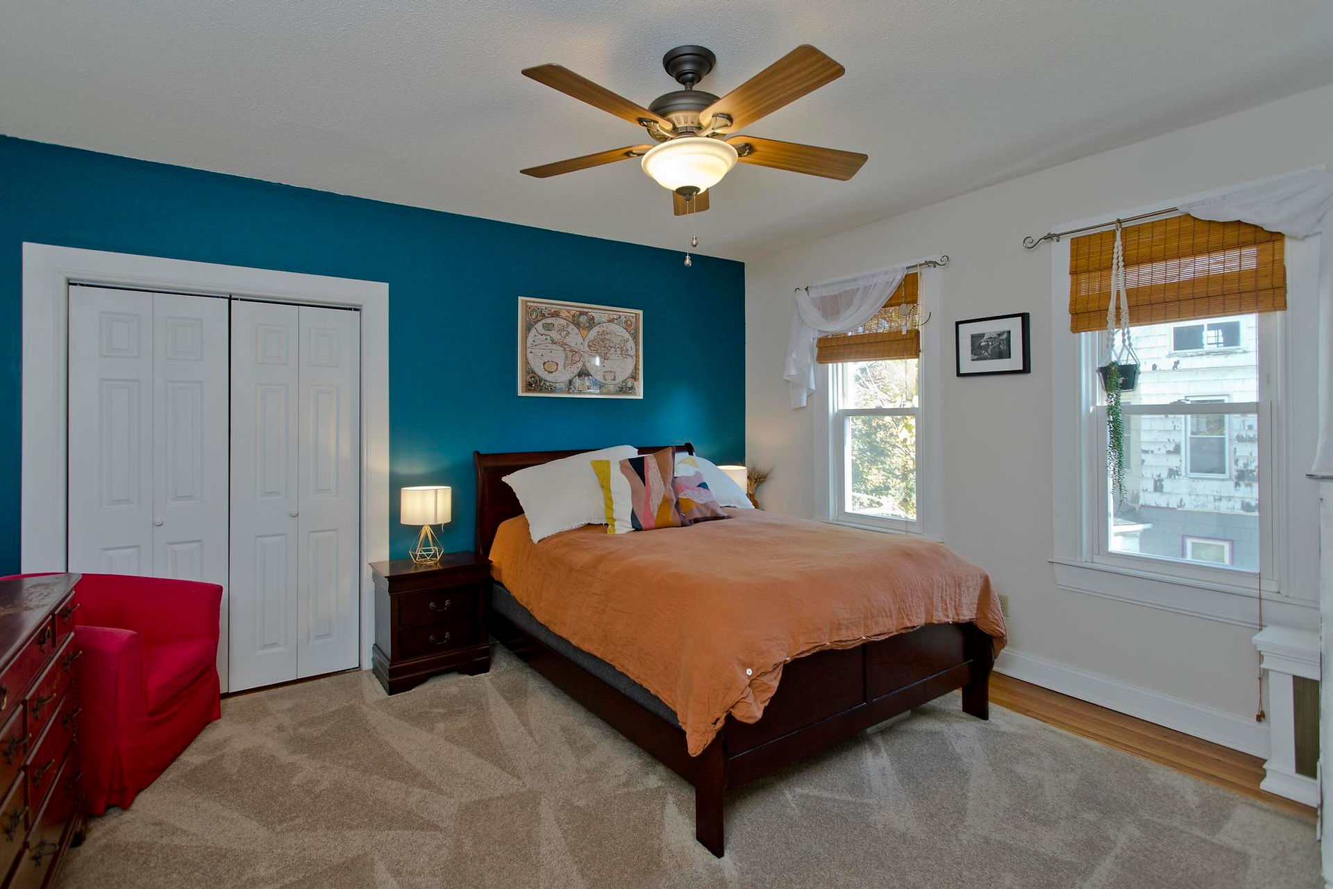 This is a good sized bedroom, with double white bifold doors on the closet, a dresser to the left next to a red accent chair, and a bed next to the wall with a window. The walls are light tan, and one accent wall is a teal color. 