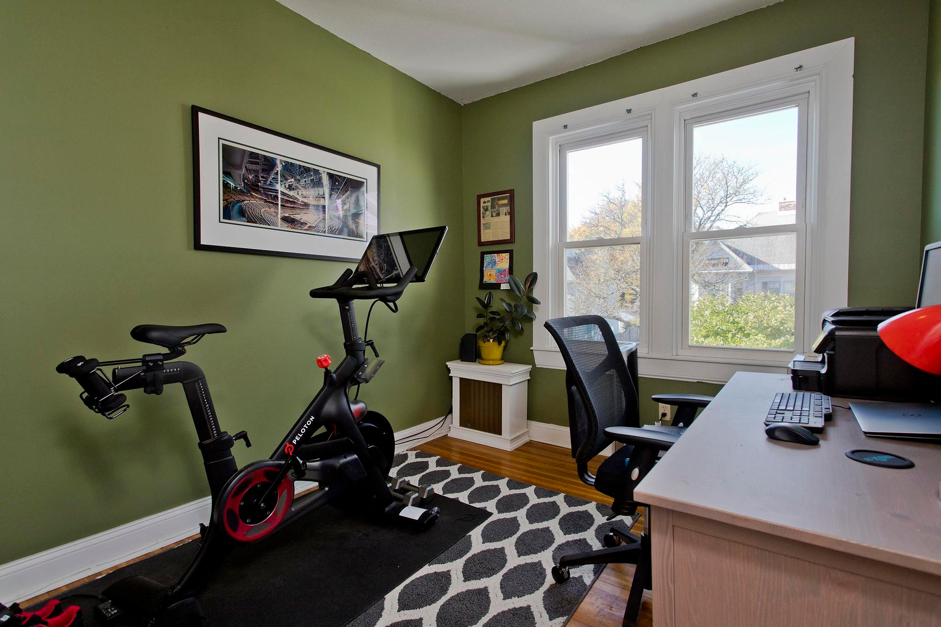 This room is green with white trim, two windows to outside sit opposite the workout bike in the room. Next to the bike there is a desk and chair.