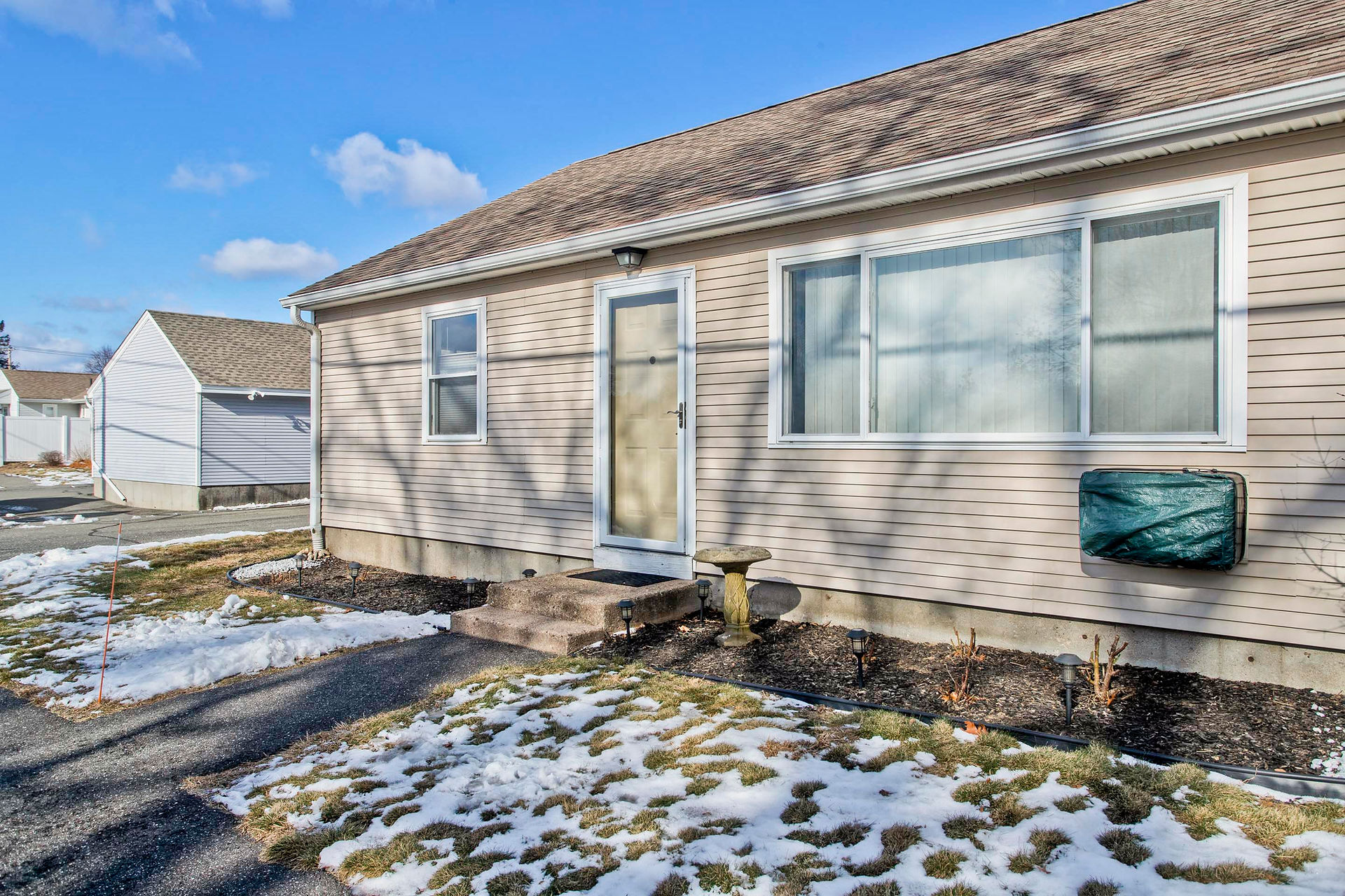The front of this 2-bedroom tan sided condo, has a large window, a front door and smll front yard, broken up in the middle by a paved walkway leading from the paved sidewalk to the door.