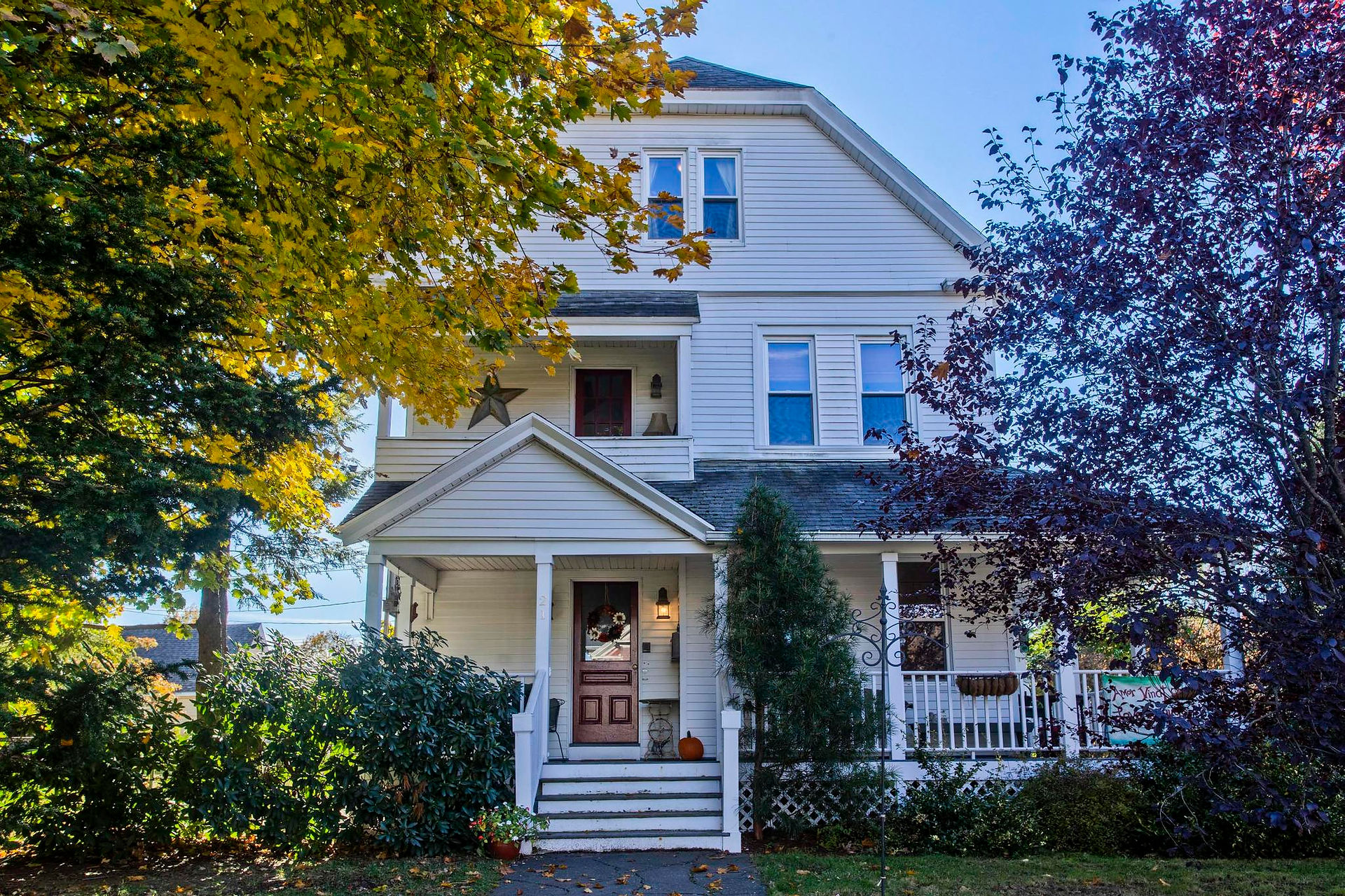 There is a white 3 story house, with a small tree in the front right yard, and a larger tree to the left. A sidewalk leads to the front door.