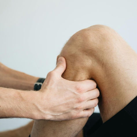 Professional performing manual knee assessment. The patient wears black shorts with a white logo. The setting is bright with a neutral background.