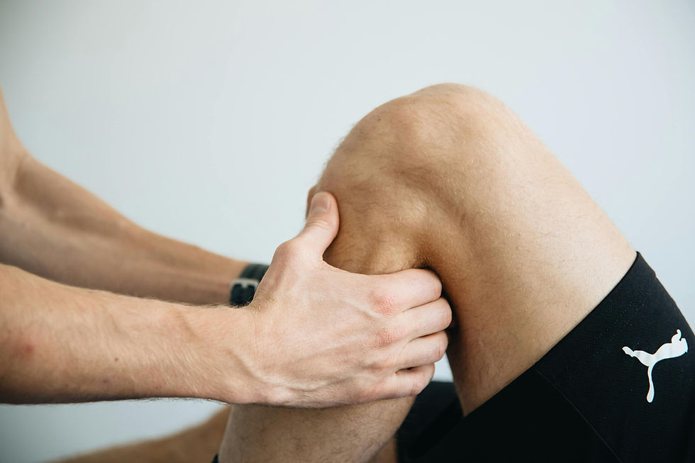 Professional performing manual knee assessment. The patient wears black shorts with a white logo. The setting is bright with a neutral background.
