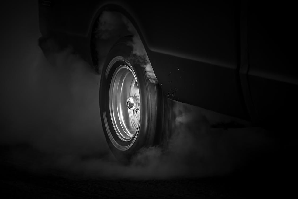 Close-up of a car tire spinning, emitting smoke in a dramatic black and white setting, suggesting high speed or burnout.