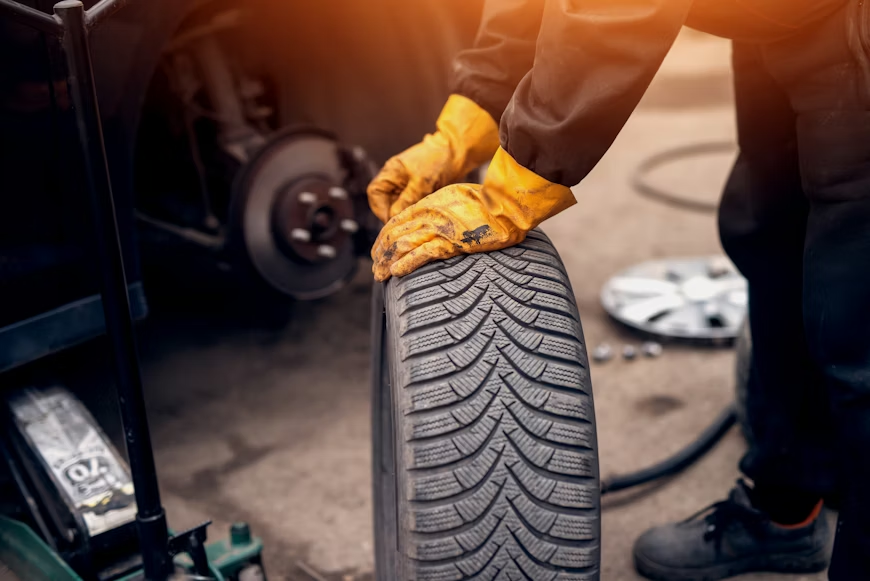 Mechanic in yellow gloves gripping a tire beside a car with removed wheel parts. Sunlight creates a warm atmosphere in the garage setting.