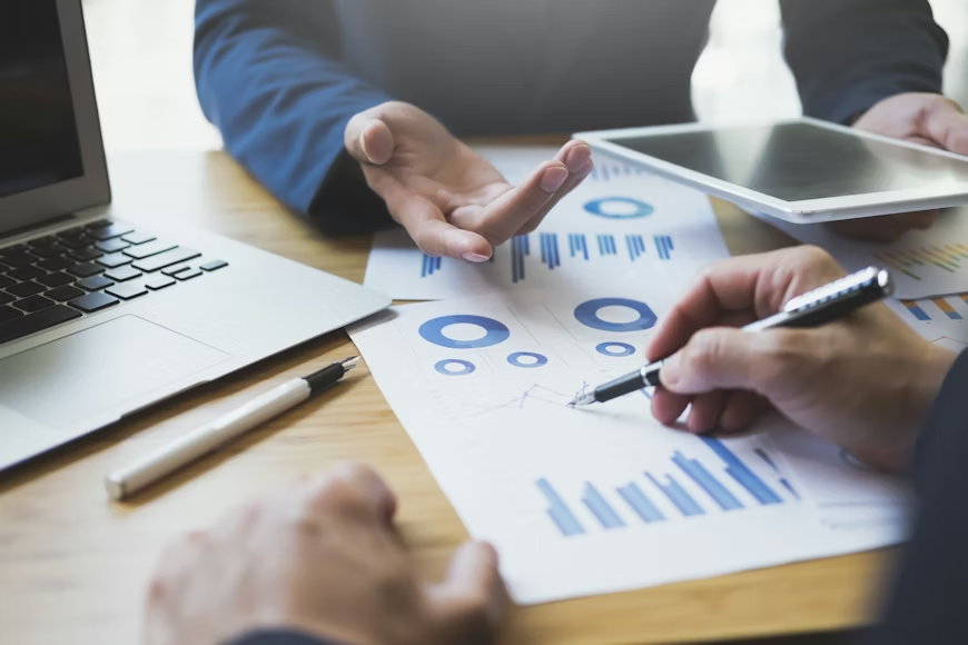 Hands discussing charts with a pen, tablet, and laptop on a wooden table. Blue graphs and diagrams suggest a professional meeting.