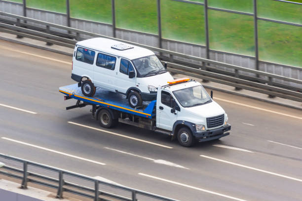 White flatbed tow truck carrying a white van on a highway, with guardrails and green grass in the background.