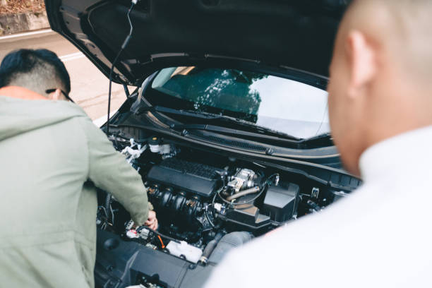 Two men inspect a car engine with the hood open, one leaning in to work, in a roadside setting.