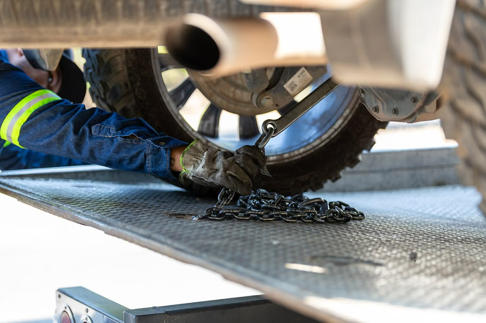 Gloved worker secures a chain under a truck tire on a flatbed trailer, with blue denim sleeve and metal deck visible.
