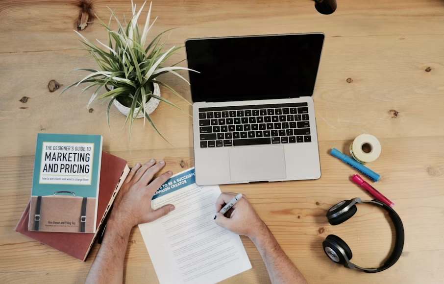 Overhead view of hands filling out a form on a wooden desk with laptop, plant, books, headphones, and colorful pens.