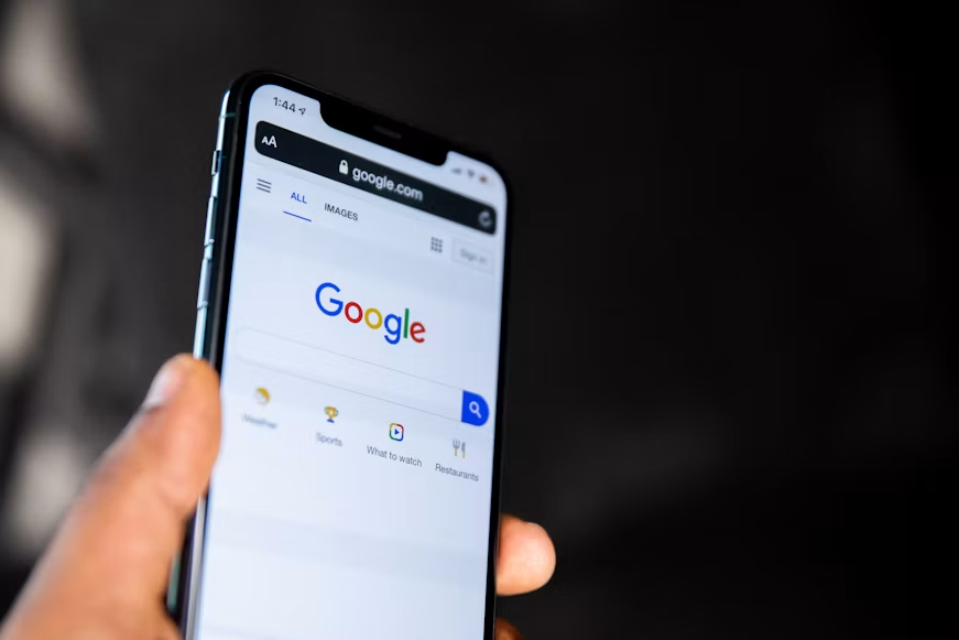 Hand holding smartphone displaying Google homepage in browser against a dark background.