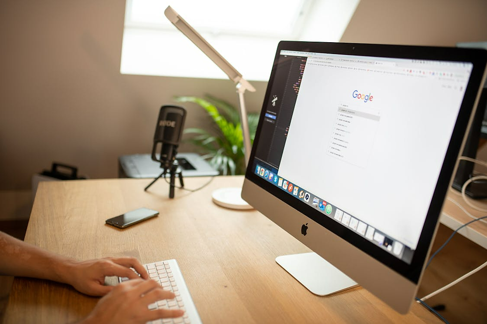 Person typing at a wooden desk with an iMac displaying Google. A microphone, smartphone, and lamp are nearby. Bright, modern setting.