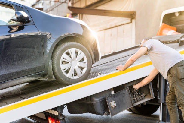 Man in a gray shirt pushes a black car onto a tow truck ramp in bright sunlight.