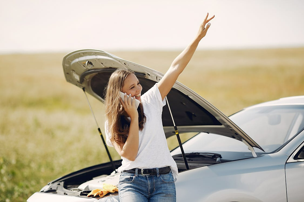 Woman stands by a broken-down car with open hood, talking on the phone and gesturing for help in a sunny field. Casual mood.